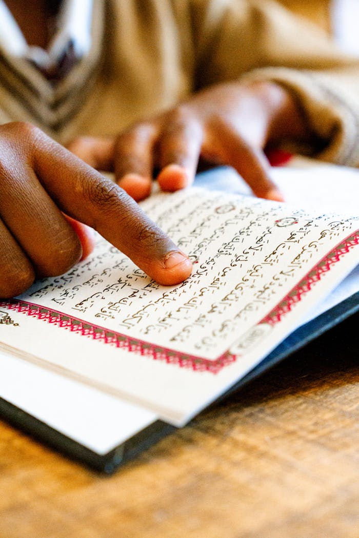 cta-02 Close-up of a child's hand pointing while reading the Quran indoors.
