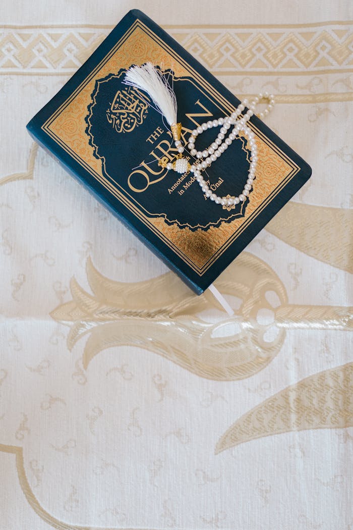 programs-01 Top view of an elegant Quran and prayer beads on ornate white cloth, symbolizing Islamic faith.